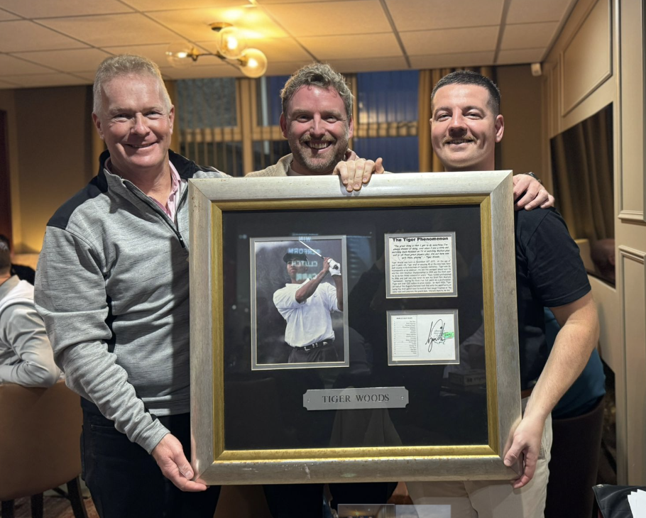Three men in a golf club clubhouse hold up a framed picture of Tiger Woods.