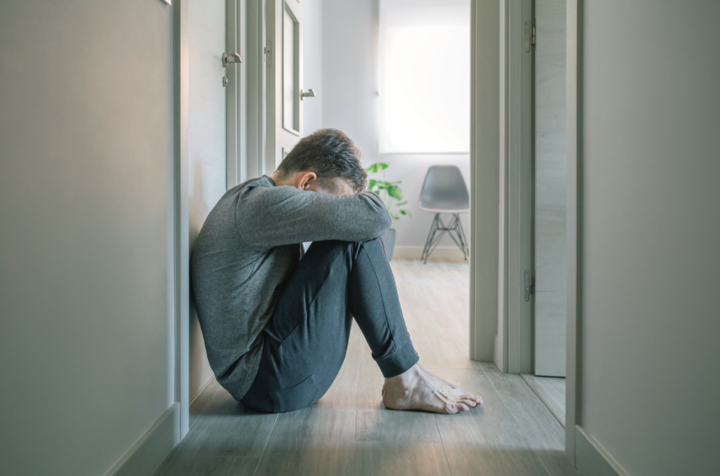 A man dressed in grey clothes crouches down in a hallway looking upset