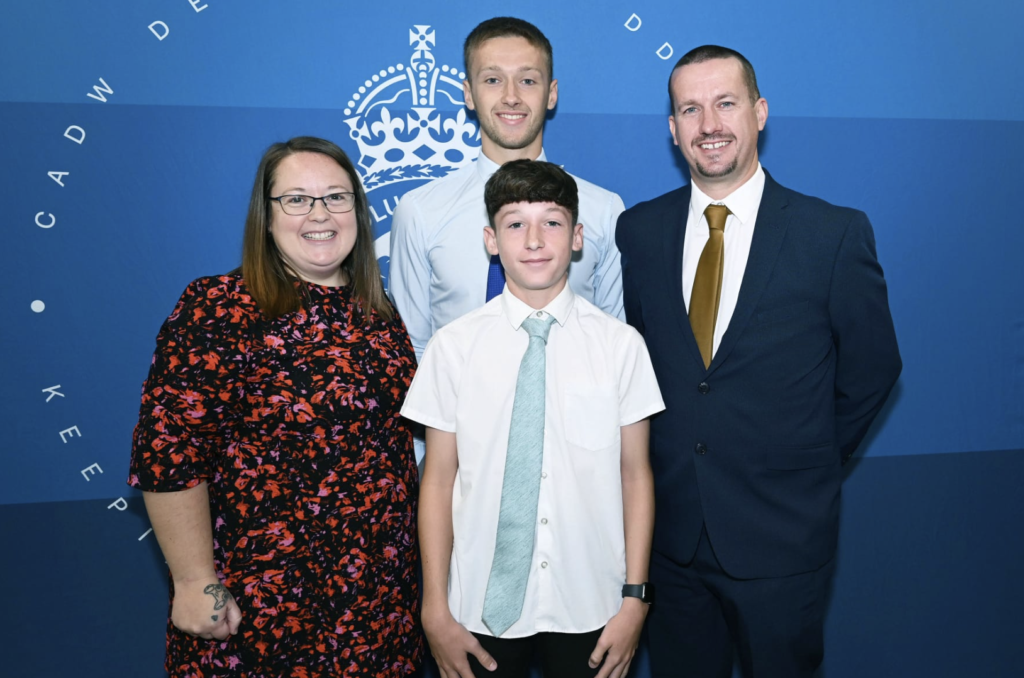 Reggie with his wife Sarah and their two children, Ceiron and Ioan smile for the camera dressed smartly in front of a blue backdrop.