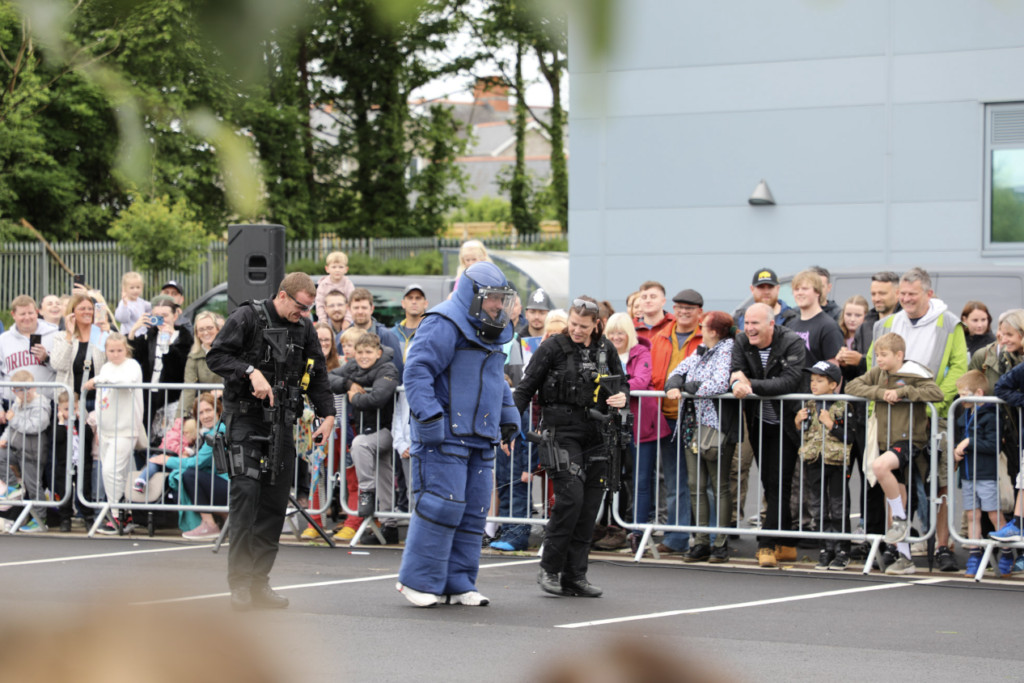 A police officer in a blue bomb disposal outfit is flanked by two officers in uniform while members of the public look on at the 2024 South Wales Police Family Fun Day